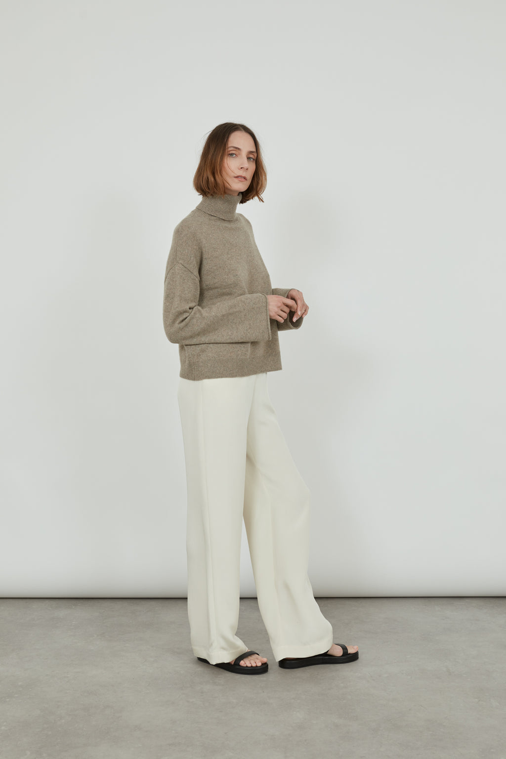 Woman is wearing the biscuit cashmere wool knit sweater, with offwhite trousers and black sandals, standing sideways.