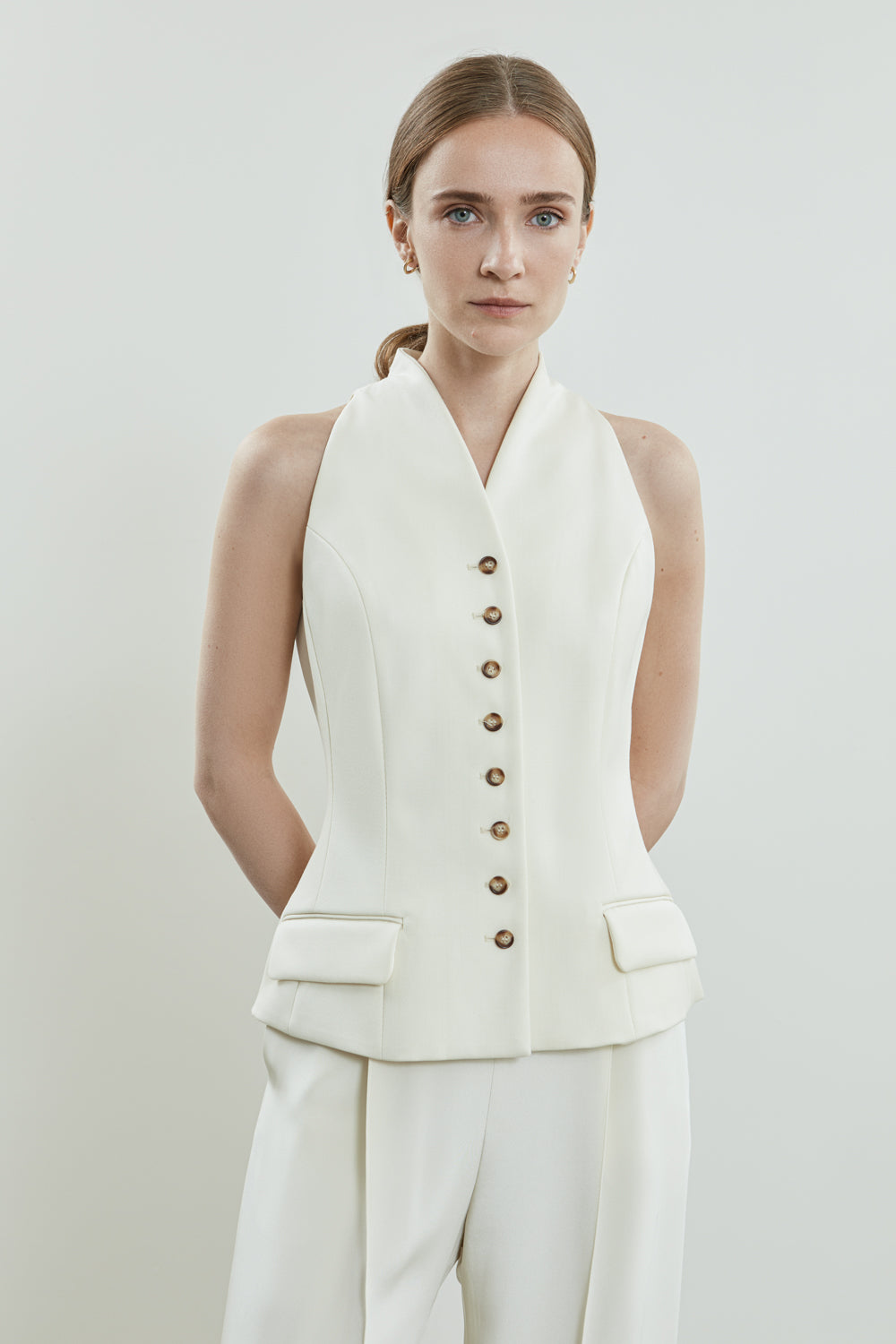 Full-length view of the off-white Akari gilet, featuring a sharp neckline, accentuated button details, and discreet flap pockets, offering a tailored.