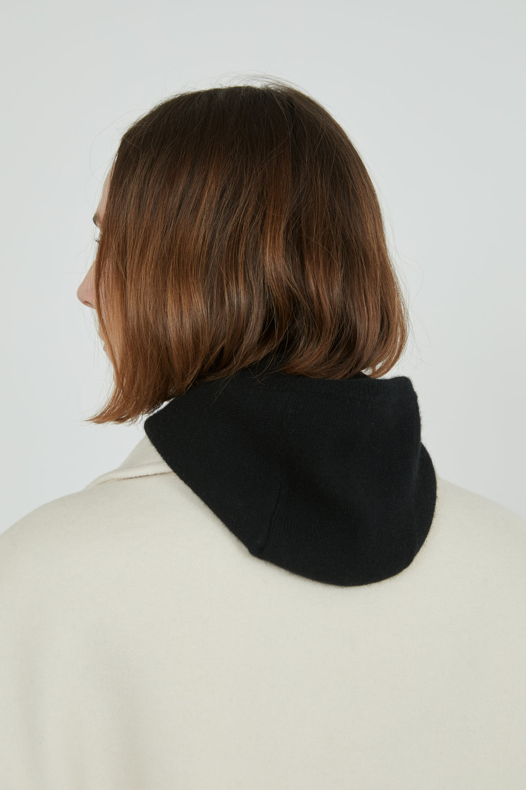 Black Cathy balaclava in merino wool combined with an offwhite cashmere coat, viewed from the back, hood off.