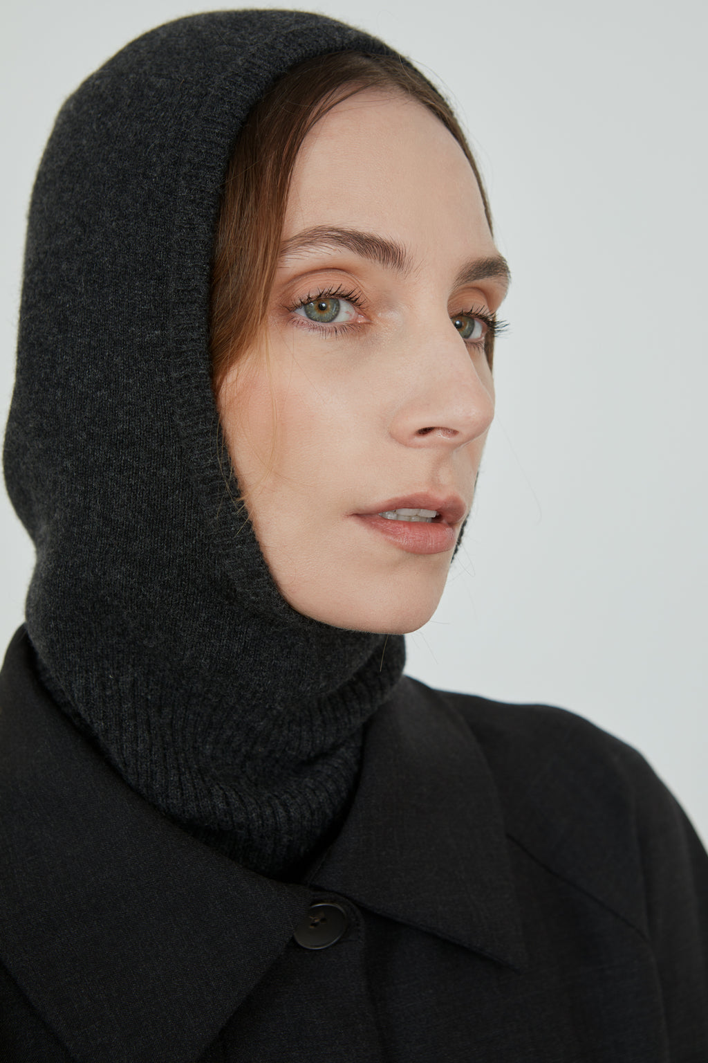 Dark grey Cathy balaclava in merino wool combined with a dark grey coat, turned sideways.
