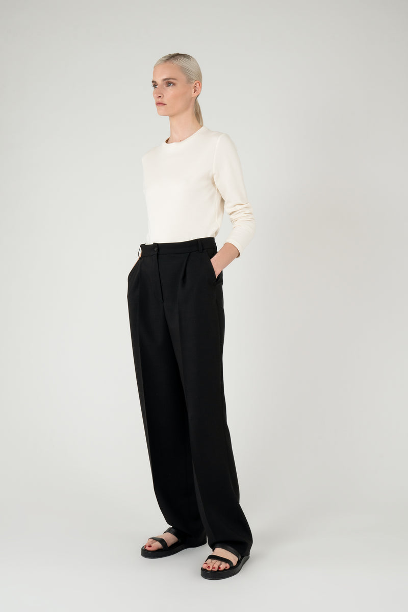 Woman wearing an off white longsleeved top with black trousers and black leather sandals, looking sideways.