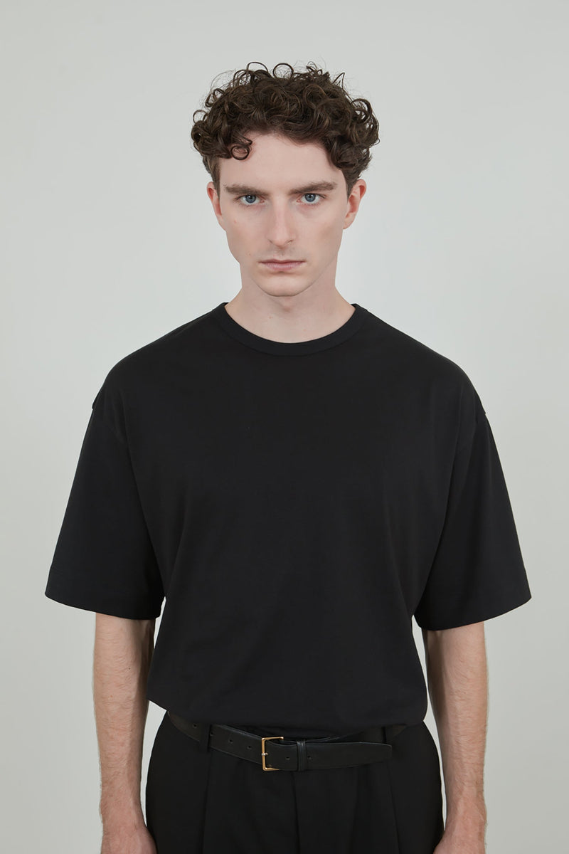 Detailed view of a man wearing a black cotton jersey T-shirt with a black leather belt and black virgin wool pants. 