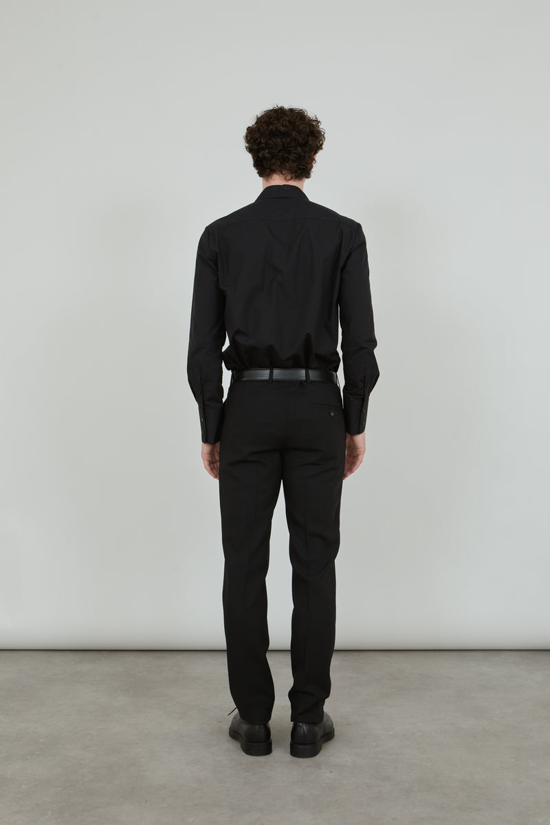 Man wearing a classic black trousers in virgin wool, combined with a classic black shirt and a black leather belt, standing with his back to the camera.. 