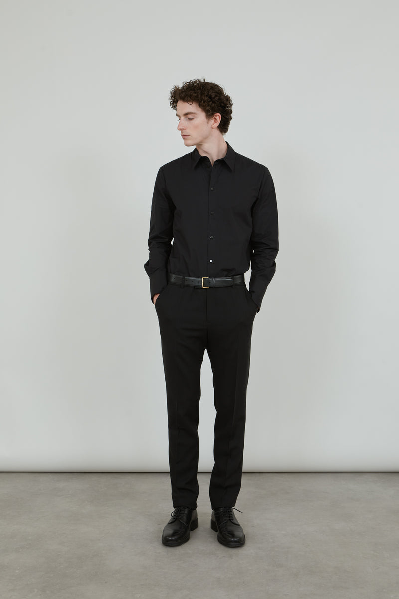 Man wearing a classic black trousers in virgin wool, combined with a classic black shirt and a black leather belt, looking sideways. 
