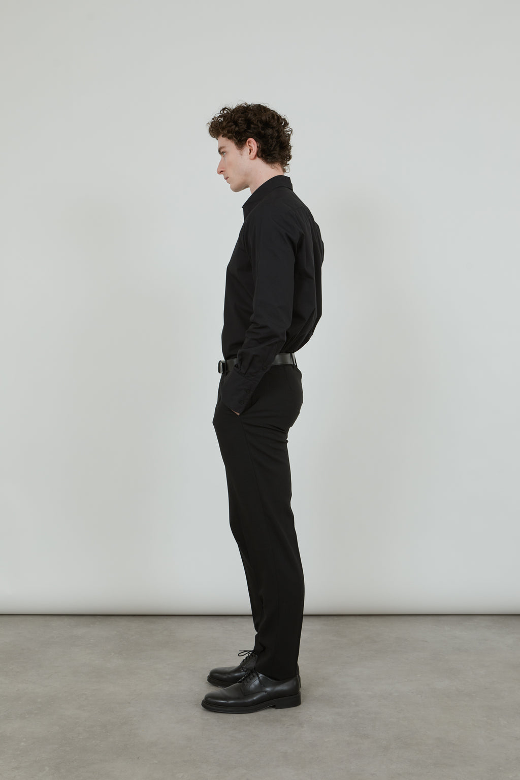 Man wearing a classic black trousers in virgin wool, combined with a classic black shirt and a black leather belt and black leather shoes,turned sideways.
