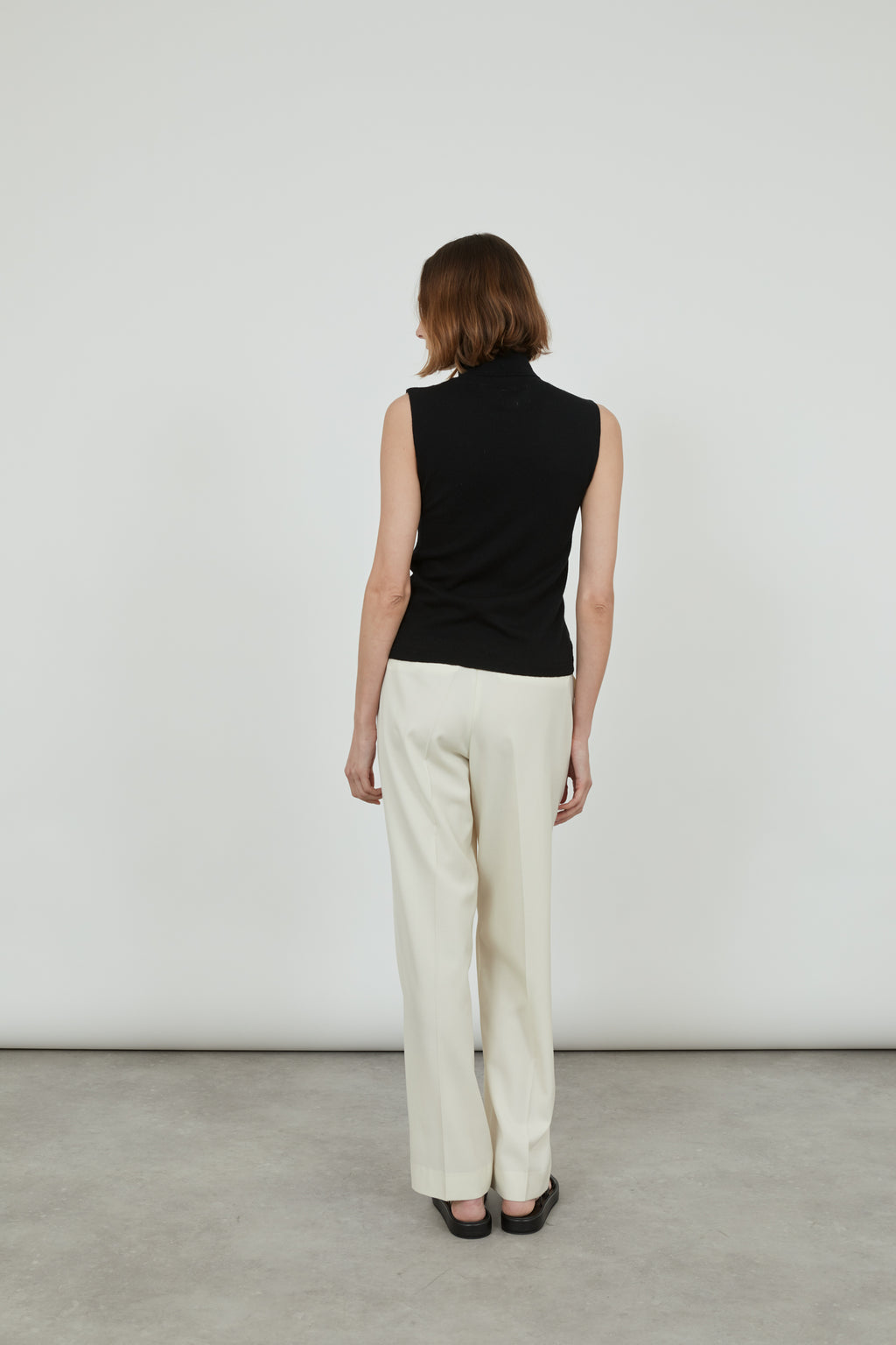Woman wearing a sleeveless black Deborah knit top in merino wool with classic offwhite pants and black leather sandals, standing with her back to the camera. . 