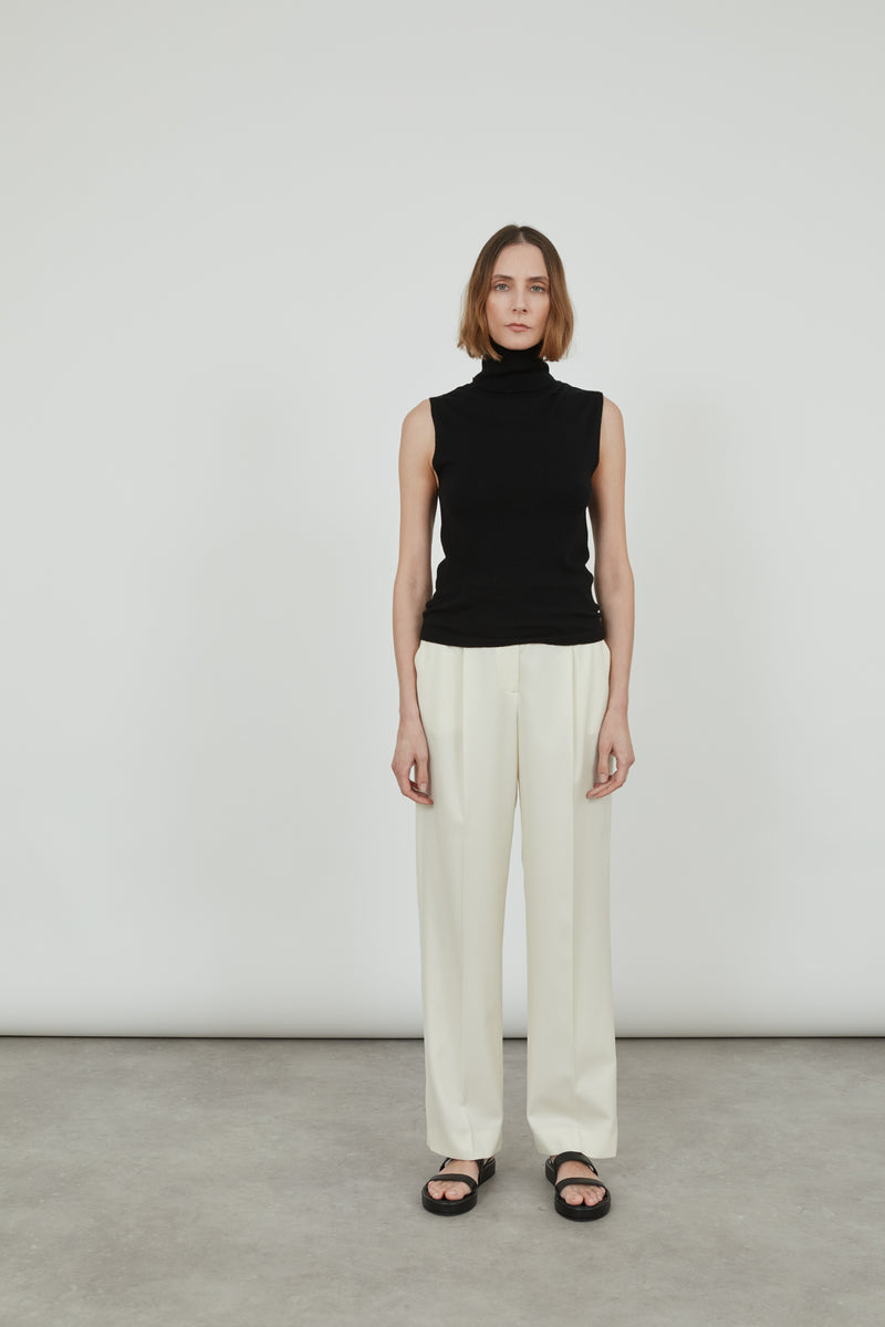 Woman wearing a sleeveless black Deborah knit top in merino wool with classic offwhite pants and black leather sandals. 