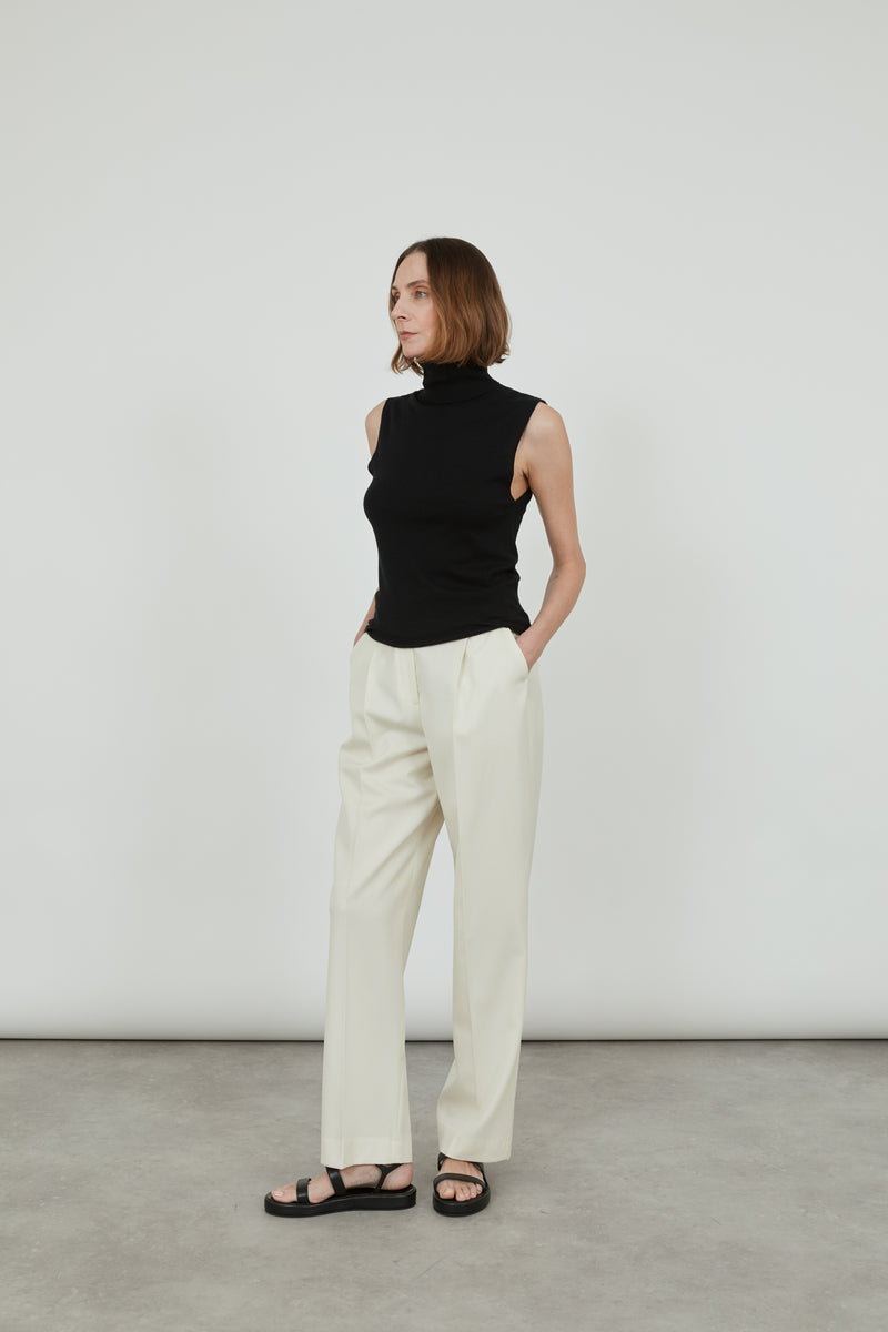Woman wearing a sleeveless black Deborah knit top in merino wool with classic offwhite pants and black leather sandals. 