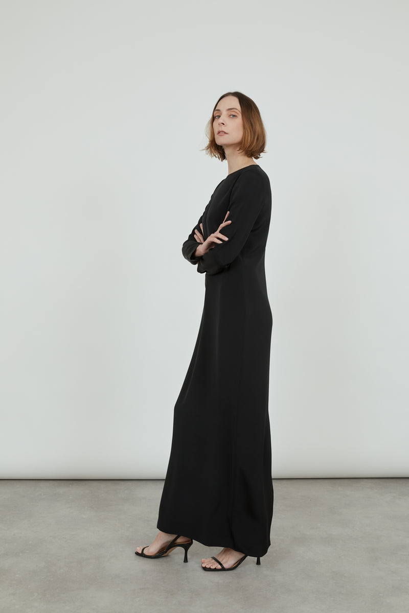 Woman wearing a long black crepe silk Abelun dress, standing sideways with her arms crossed.