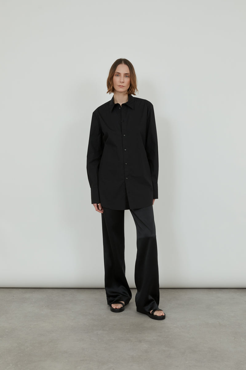 A woman is wearing a black, cotton poplin Adam shirt with a black satin trousers.