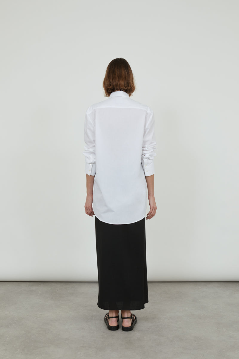 Person wearing a classic white Adam shirt in cotton poplin, with a long black skirt and black leather sandals, standing with the back to the camera.