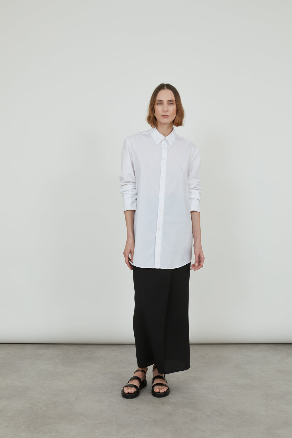 Person wearing a classic white Adam shirt in cotton poplin, with a long black skirt and black leather sandals.. 