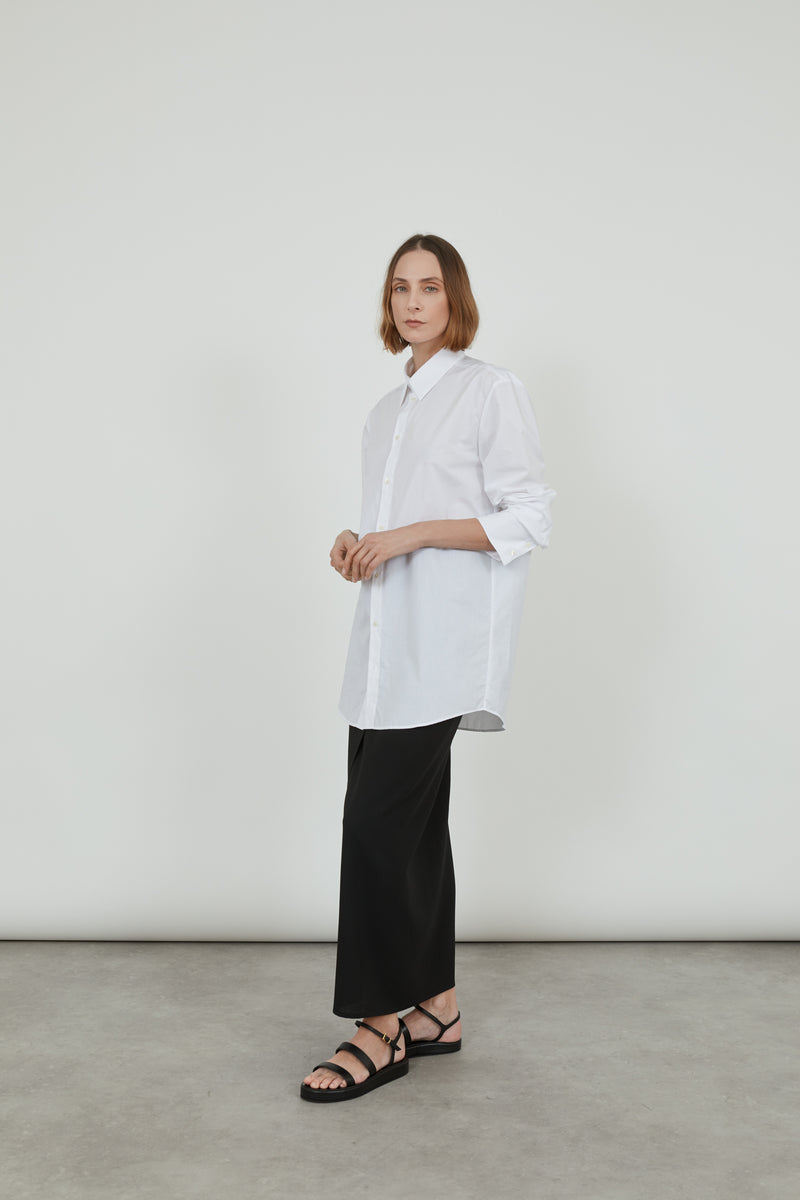 Person wearing a classic white Adam shirt in cotton poplin, with a long black skirt and black leather sandals, standing sideways.