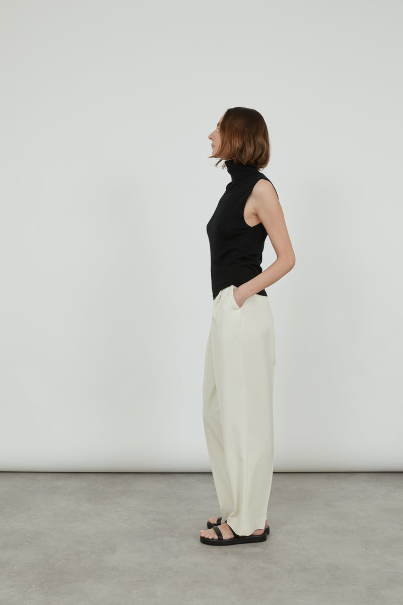 Woman wearing a sleeveless black Deborah knit top in merino wool with classic offwhite pants and black leather sandals, standing sideways.
