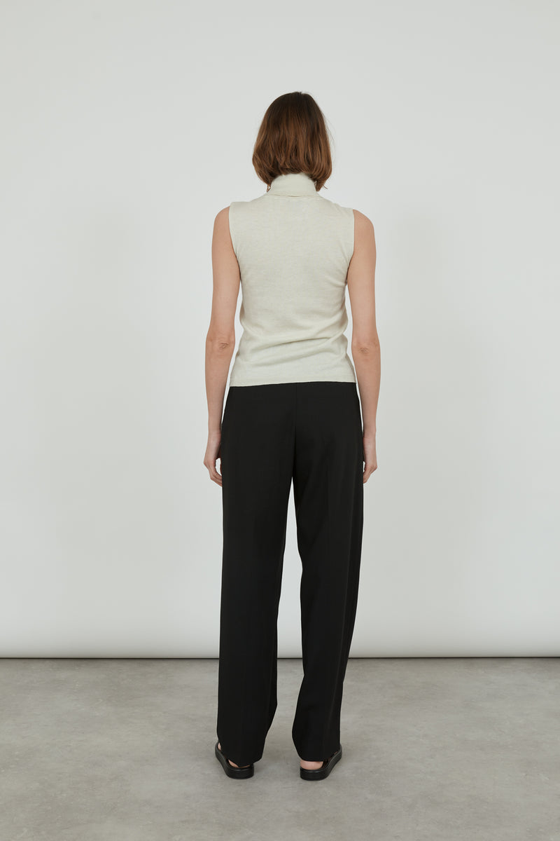 Woman wearing a sleeveless offwhite Deborah knitted turtleneck top in merino wool with classic black pants and black leather sandals, standing with her back to the camera.