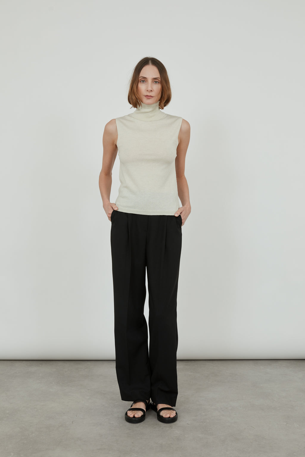 Woman wearing a sleeveless offwhite Deborah knitted top in merino wool with classic black pants and black leather sandals, standing with her hands in her pockets.. 