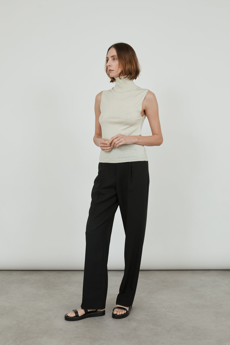 Woman wearing a sleeveless offwhite Deborah knitted turtleneck top in merino wool with classic black pants and black leather sandals, standing sideways.