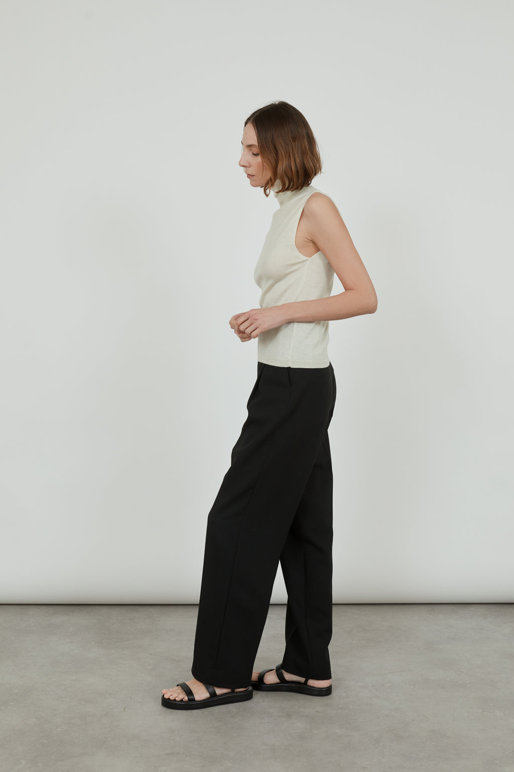 Woman wearing a sleeveless offwhite Deborah knitted turtleneck top in merino wool with classic black pants and black leather sandals, turned to the right.
