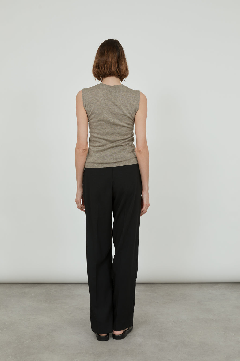 Woman wearing a sleeveless biscuit Emma knit top in merino wool with classic black pants and black leather sandals, standing with her back to the camera.. 