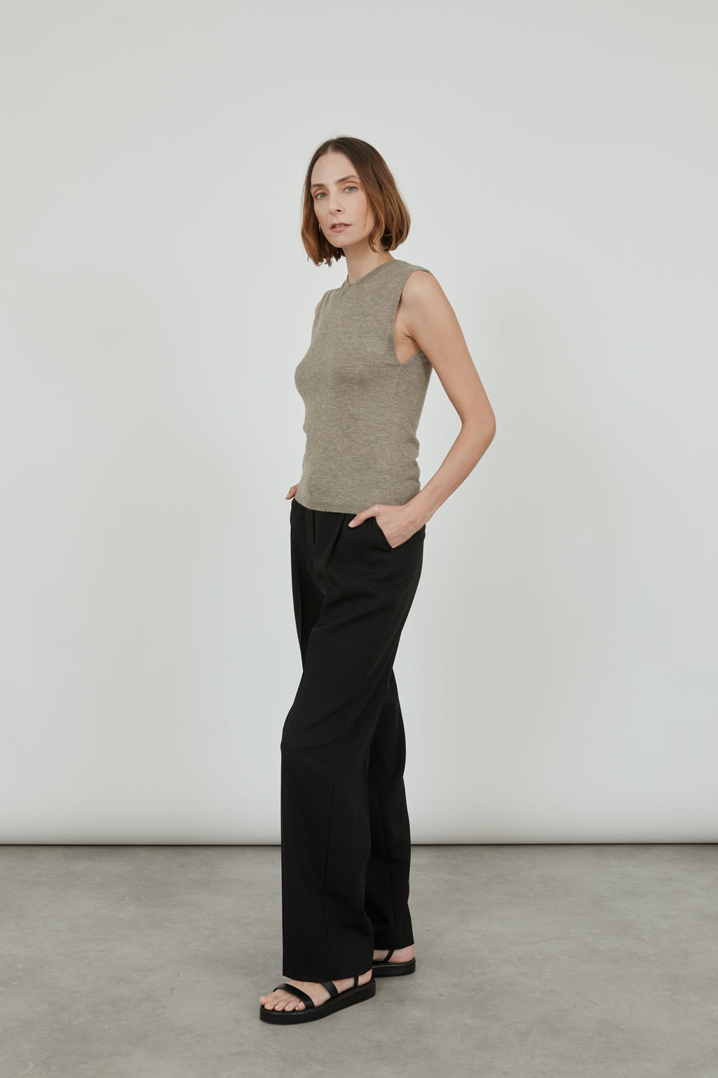 Woman wearing a sleeveless biscuit Emma knit top in merino wool with classic black pants and black leather sandals, standing sideways. 
